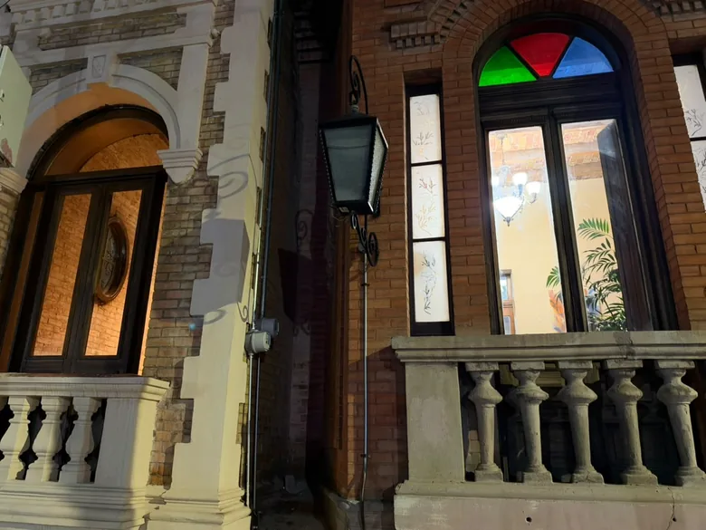 Detalle de la fachada de noche: farol de herrería, balaustrada de cantera y un vitral de colores sobre la ventana.