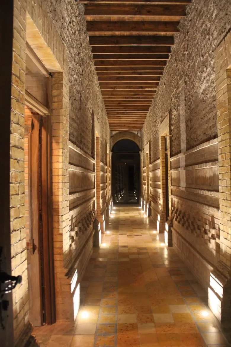 Corredor largo de la planta baja, con techo de vigas de madera, muros de piedra y ladrillo y luces a ras de piso.